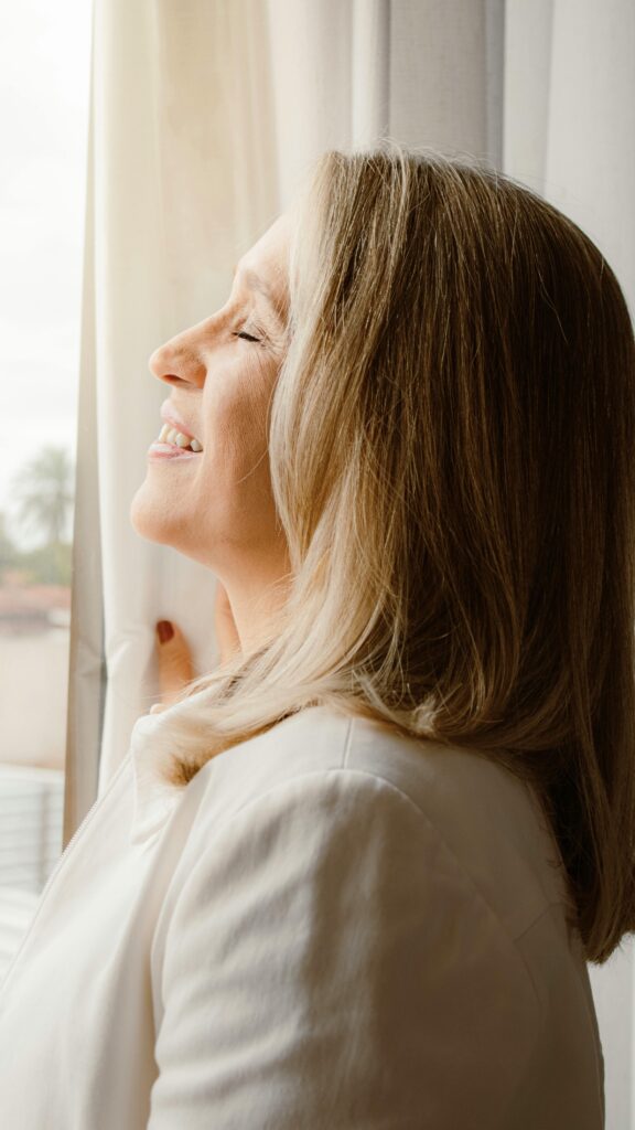 A senior woman enjoys a peaceful moment with eyes closed by a sunlit window indoors.